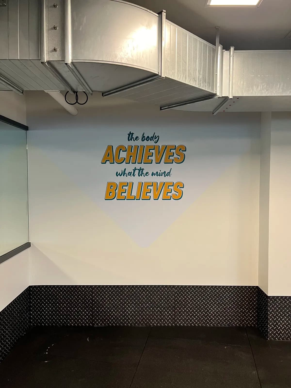 Vægfolie med teksten 'The body achieves what the mind believes' i fitnesscenter