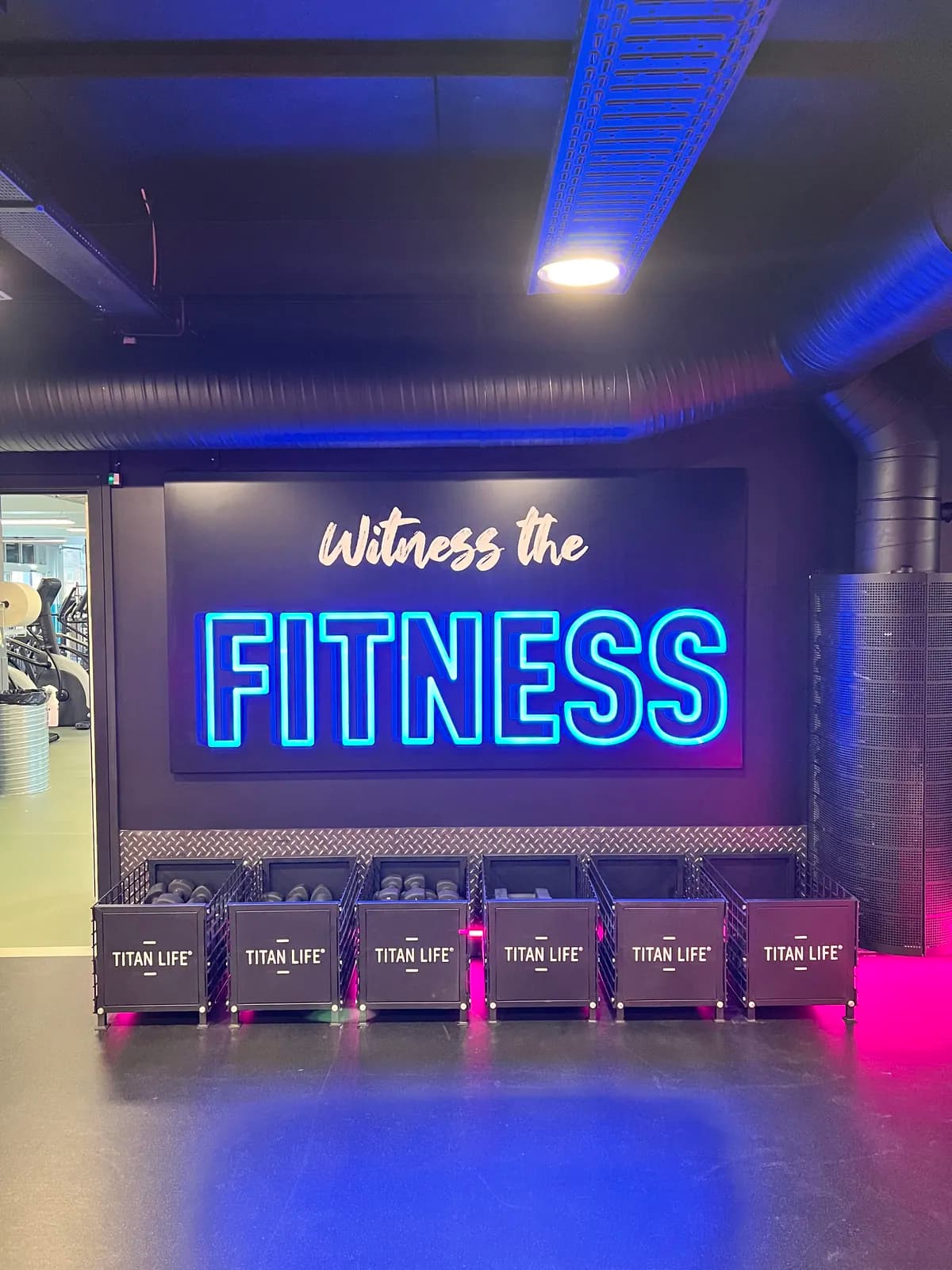 LED-neonskilt med teksten 'Witness the fitness' i blåt i mørklagt fitnessområde