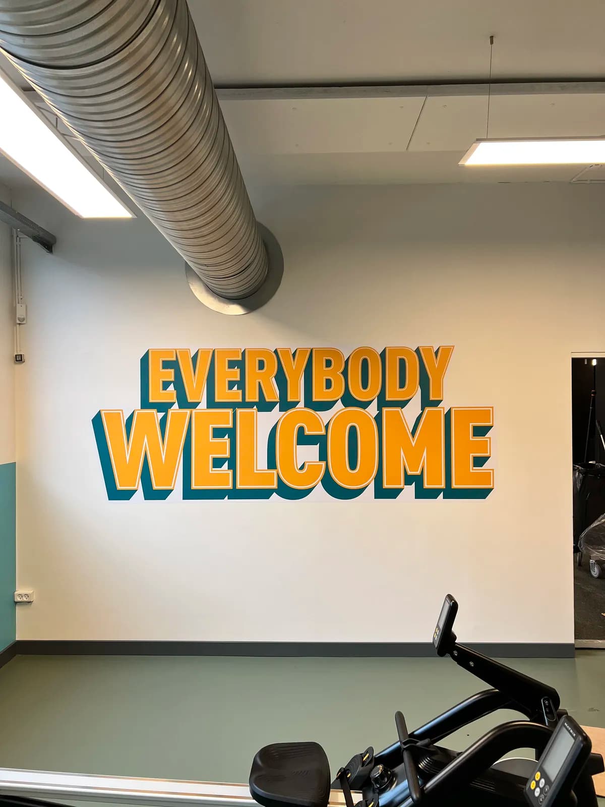 Vægfolie med teksten 'Everybody welcome' i gult og petroleum i fitnesscenter
