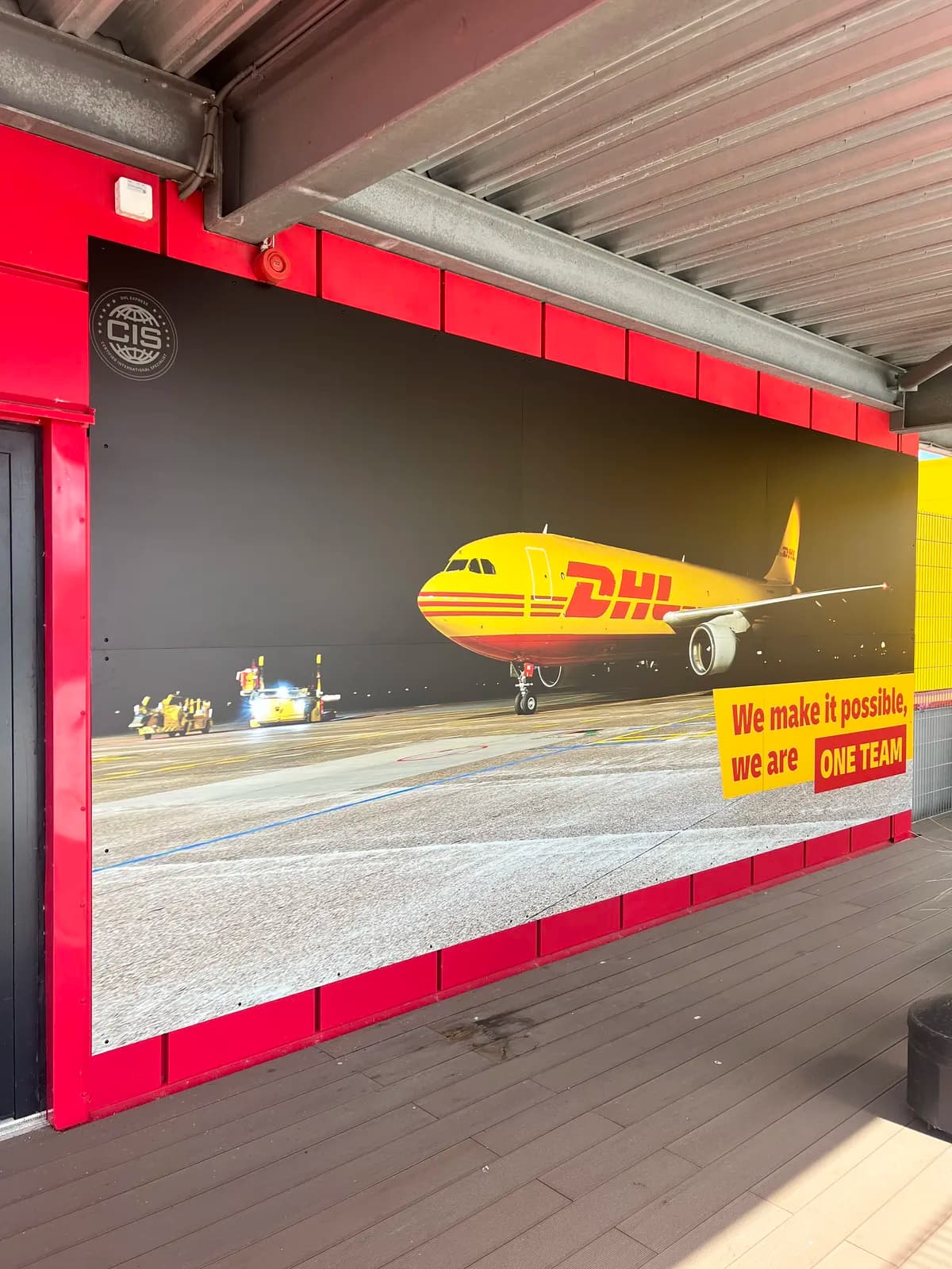 Printede facadeplader med DHL-fragtfly og teamslogan monteret på rød facade hos DHL Express